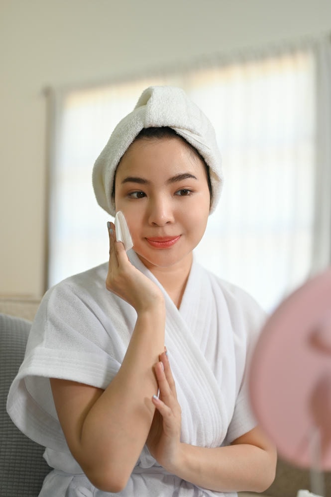 các bước skincare cơ bản. Attractive Asian woman in bathrobe cleaning her face with cleansing oil applying moisturizer