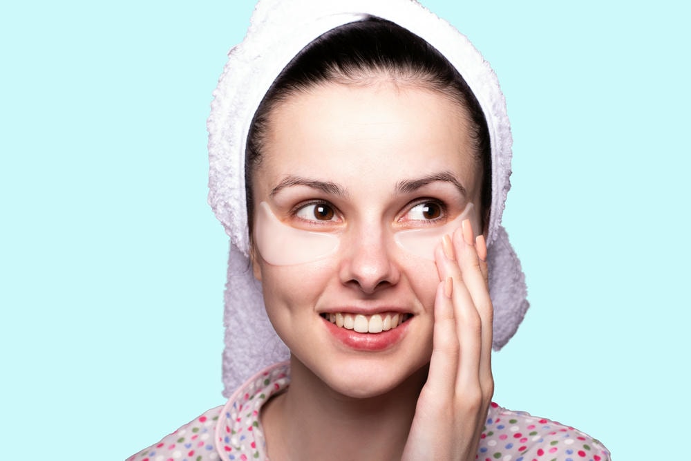 cách trị thâm mắt. Người phụ nữ trẻ tươi cười quấn khăn trên đầu và đắp mặt nạ