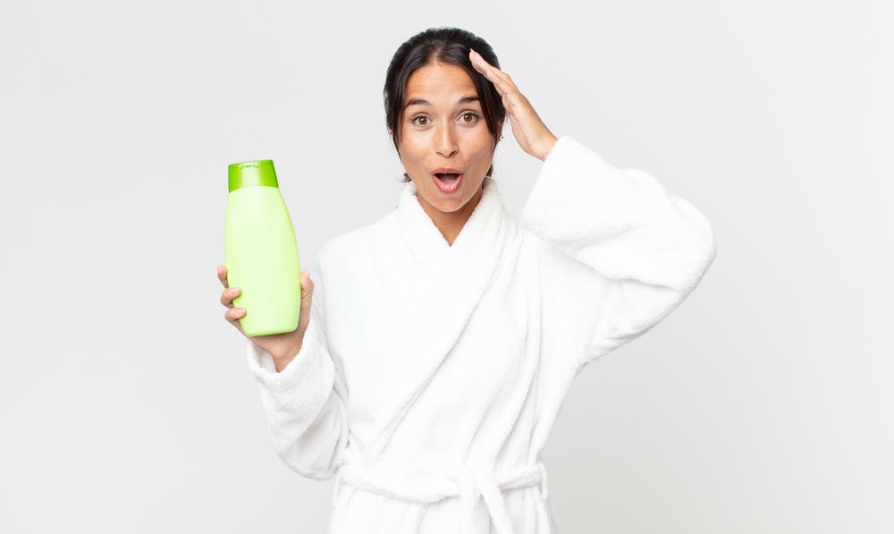 dầu gội trị gàu tốt nhất hiện nay. Người phụ nữ gốc Hispanic trẻ tuổi trông vui vẻ, kinh ngạc và ngạc nhiên khi mặc áo choàng tắm và cầm dầu gội
